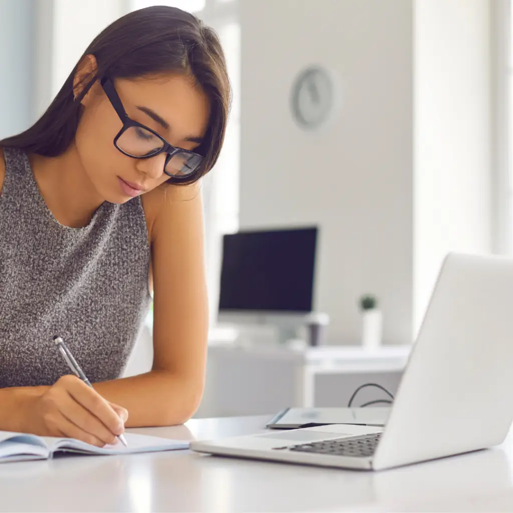 A calm woman in glasses is writing on a notebook in front of a laptop.
