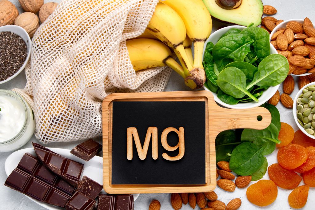 "Assorted magnesium-rich foods including bananas, spinach, almonds, pumpkin seeds, dried apricots, dark chocolate, chia seeds, avocado, and yogurt arranged around a chalkboard with ‘Mg’ symbol, highlighting natural dietary sources of magnesium.