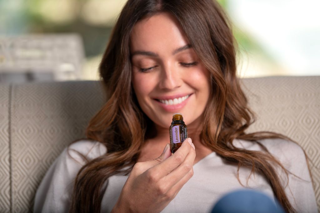 Smiling woman sitting on a couch while holding and smelling a bottle of essential oil, illustrating the calming benefits of essential oils for autism