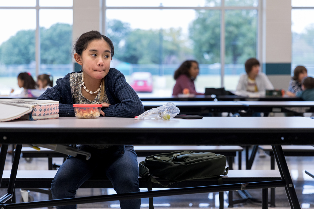 A child sitting alone in a school cafeteria, appearing disengaged, symbolizing emotion dysregulation and social challenges in kids.